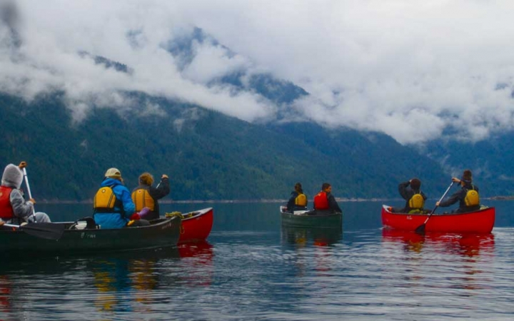 a group of outward bound students paddle canoes on calm water with foggy mountains in the background